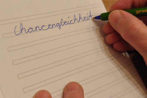 Nahaufnahme des handschriftlich geschribenen Wortes "Chancengleichheit" in Schreibschrift auf einem doppelt-liniertem Papier. Hand und Kugelschreiber sind ebenfalls abgelichtet.